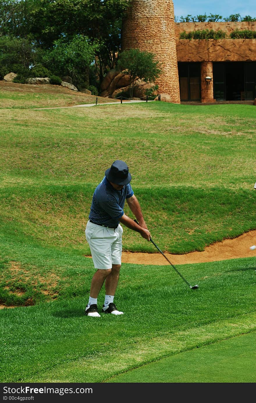 Middle age golfer hitting the ball onto the green. Halfway house in the background. Golf club is in motion. Middle age golfer hitting the ball onto the green. Halfway house in the background. Golf club is in motion.