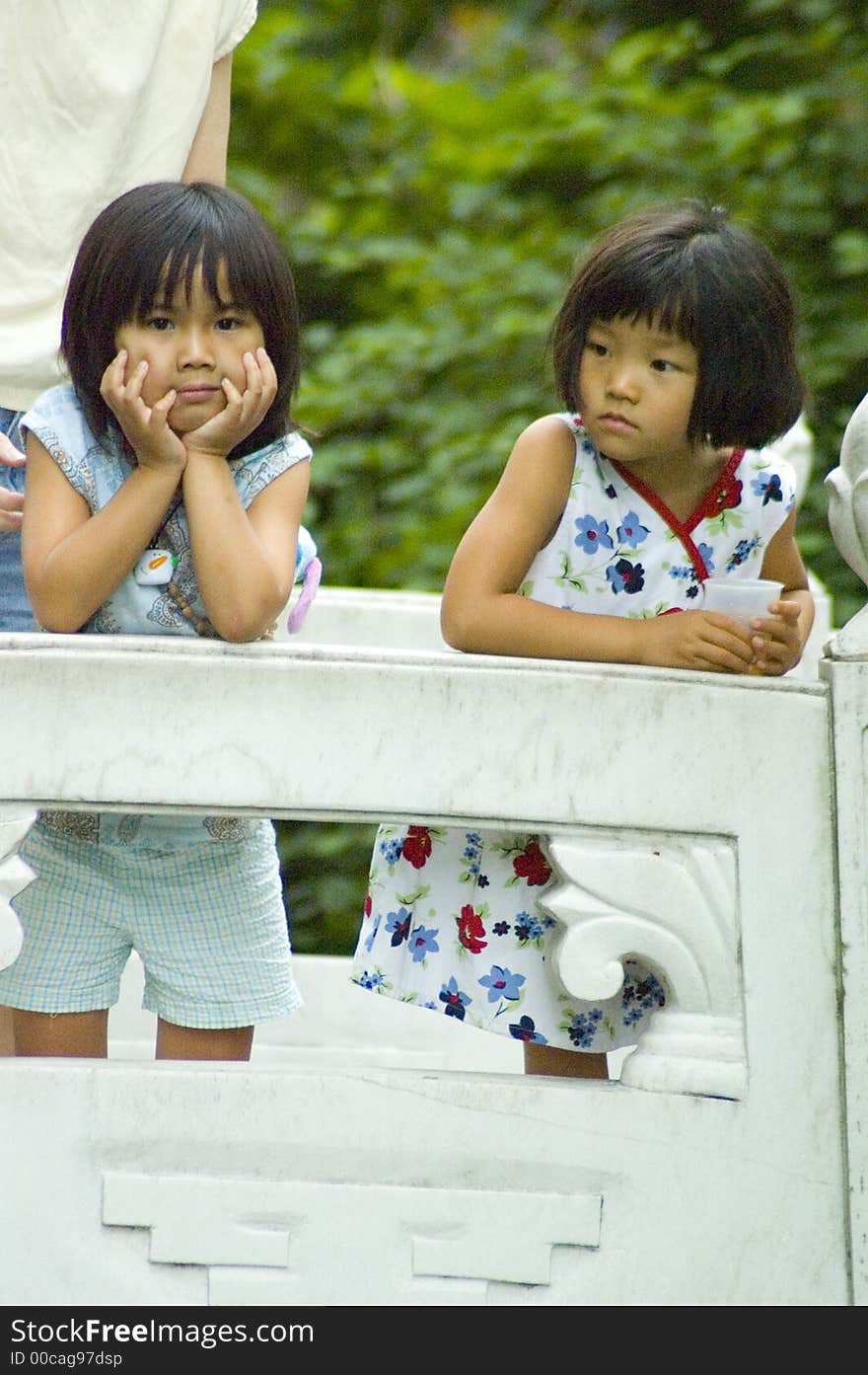Two cute young Asian girls lean on the rail of a stone bridge, one thoughtfully holding her face in her hands and looking at the camera. Two cute young Asian girls lean on the rail of a stone bridge, one thoughtfully holding her face in her hands and looking at the camera.