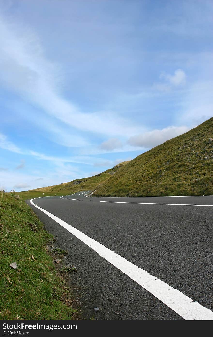 Uphill road in deserted countryside with grass verges either side with a blue sky with clouds. Set in the Brecon Beacons, National Park, Wales, United Kingdom. Uphill road in deserted countryside with grass verges either side with a blue sky with clouds. Set in the Brecon Beacons, National Park, Wales, United Kingdom.