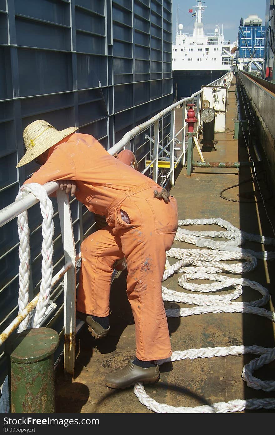 Sailor dressed in orange overall with a hat, watching the loading of a large steel module onboard a freight ship. Sailor dressed in orange overall with a hat, watching the loading of a large steel module onboard a freight ship.