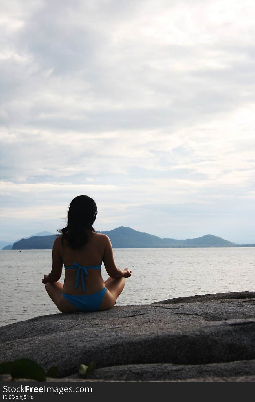 A girl in blue bikini sitted cross-legged on a rock by the beach, meditating. A girl in blue bikini sitted cross-legged on a rock by the beach, meditating.