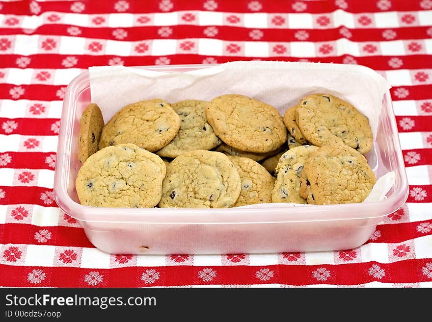 A container of homemade chocolate chip cookies on gingham tablecloth. A container of homemade chocolate chip cookies on gingham tablecloth.