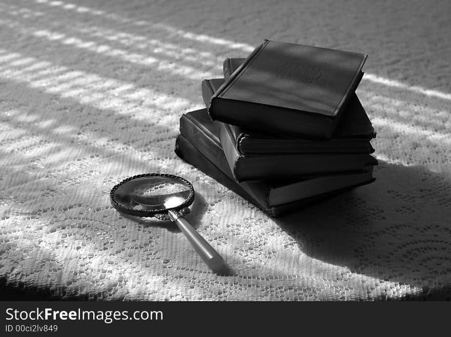 Stack of old books with reading glass on sunlit laced tabletop, black and white. Stack of old books with reading glass on sunlit laced tabletop, black and white