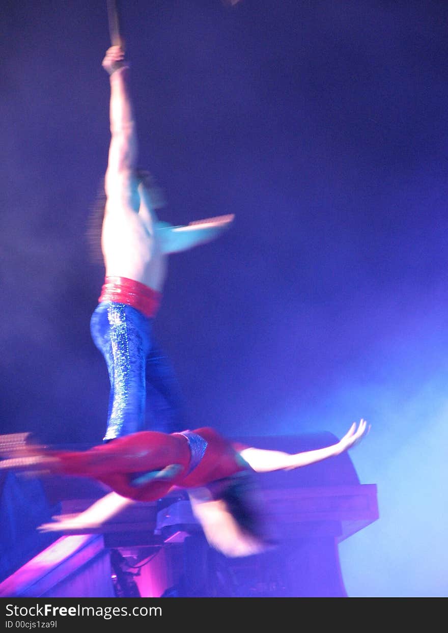 2 aerial acrobats in a spin above the stage, multi-blue/red colors with a misty blue background. Man holding on to rope with one arm as woman is at his feet horizontaly. 2 aerial acrobats in a spin above the stage, multi-blue/red colors with a misty blue background. Man holding on to rope with one arm as woman is at his feet horizontaly.