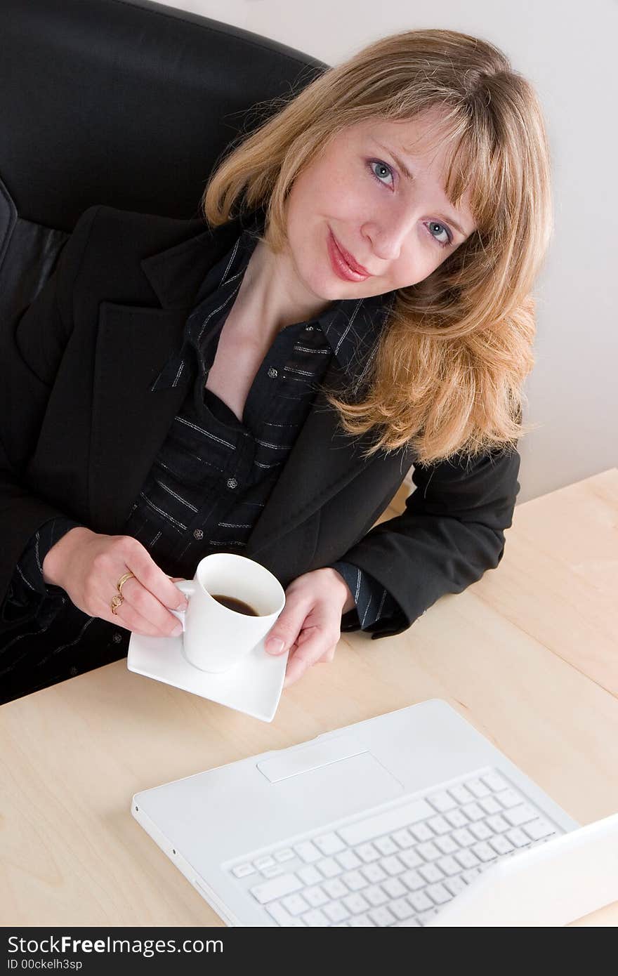 A blond woman in a black suit drinking a cup of coffee at her desk. A blond woman in a black suit drinking a cup of coffee at her desk