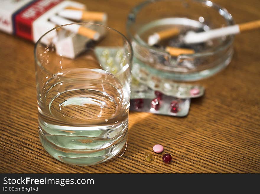 Glass with water, cigarettes, tablets and ashtray on a table, (studio). Glass with water, cigarettes, tablets and ashtray on a table, (studio).