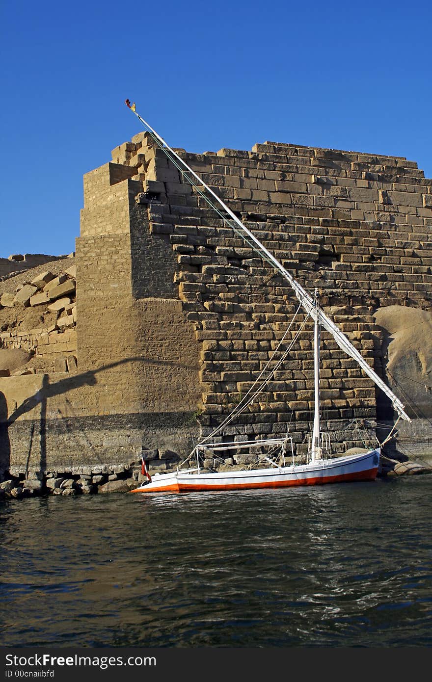 Anchored Egyptian traditional sailing boat know as Felucca shot against a beautiful blue sky on a sunny winter day in Aswan. The background is an ancient building with some phraonic scripts on some of its stones. Anchored Egyptian traditional sailing boat know as Felucca shot against a beautiful blue sky on a sunny winter day in Aswan. The background is an ancient building with some phraonic scripts on some of its stones.