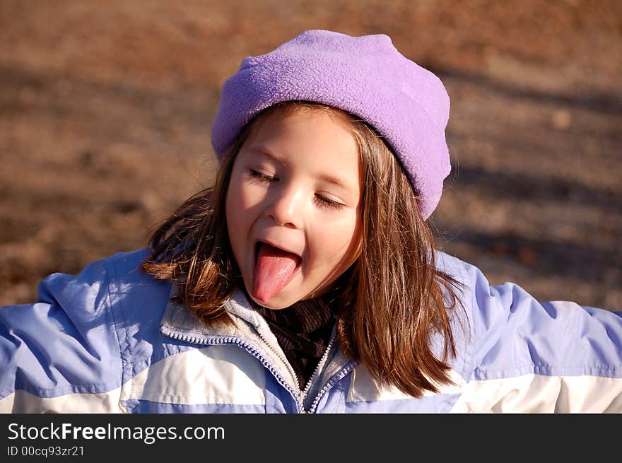 Four year old girl dressed in a purple hat and coat. She is sticking out her tongue with her arms stretched out and the late autumn sun is on her face. Four year old girl dressed in a purple hat and coat. She is sticking out her tongue with her arms stretched out and the late autumn sun is on her face.