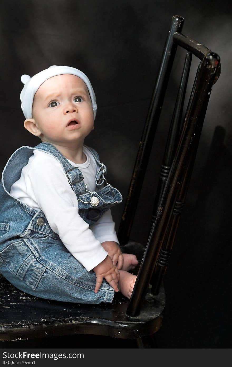 Image of adorable baby wearing denim overalls sitting on a black chair. Image of adorable baby wearing denim overalls sitting on a black chair