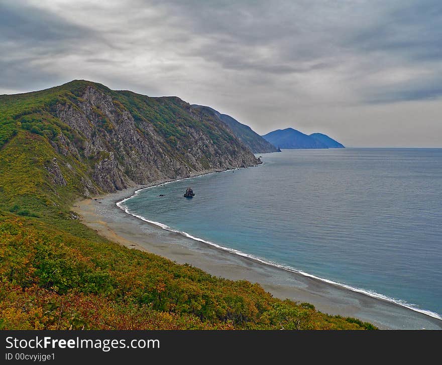 The dark clouds on whole sky above the capes and sea. Dusk. Autumn. The yellow bushes and grey beach with line of white surf are on foreground. Japanese sea, Russian Far East, Primorye, Tasovaya bay. The dark clouds on whole sky above the capes and sea. Dusk. Autumn. The yellow bushes and grey beach with line of white surf are on foreground. Japanese sea, Russian Far East, Primorye, Tasovaya bay.