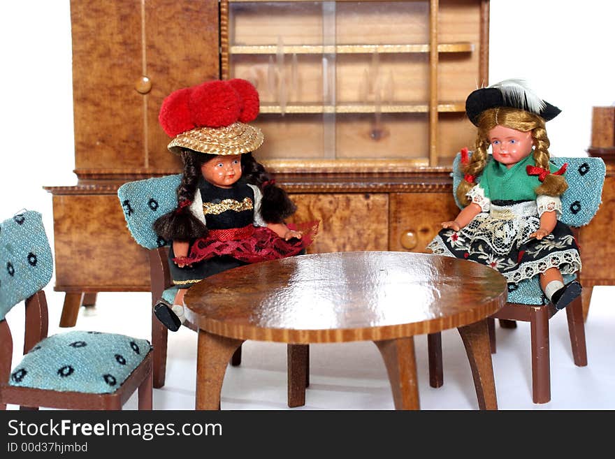 2 dolls in traditional European (I think they are Austrian but not sure) dresses sitting on chairs round a table (old hand made furniture) with racks on the background. 2 dolls in traditional European (I think they are Austrian but not sure) dresses sitting on chairs round a table (old hand made furniture) with racks on the background.