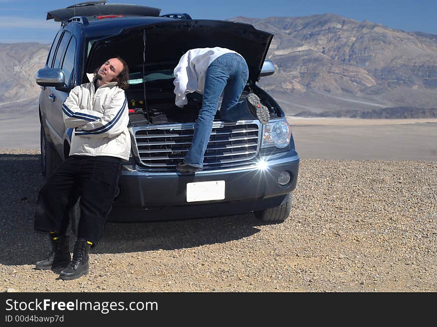 Woman Trying To Fix Car While Husband Relaxes