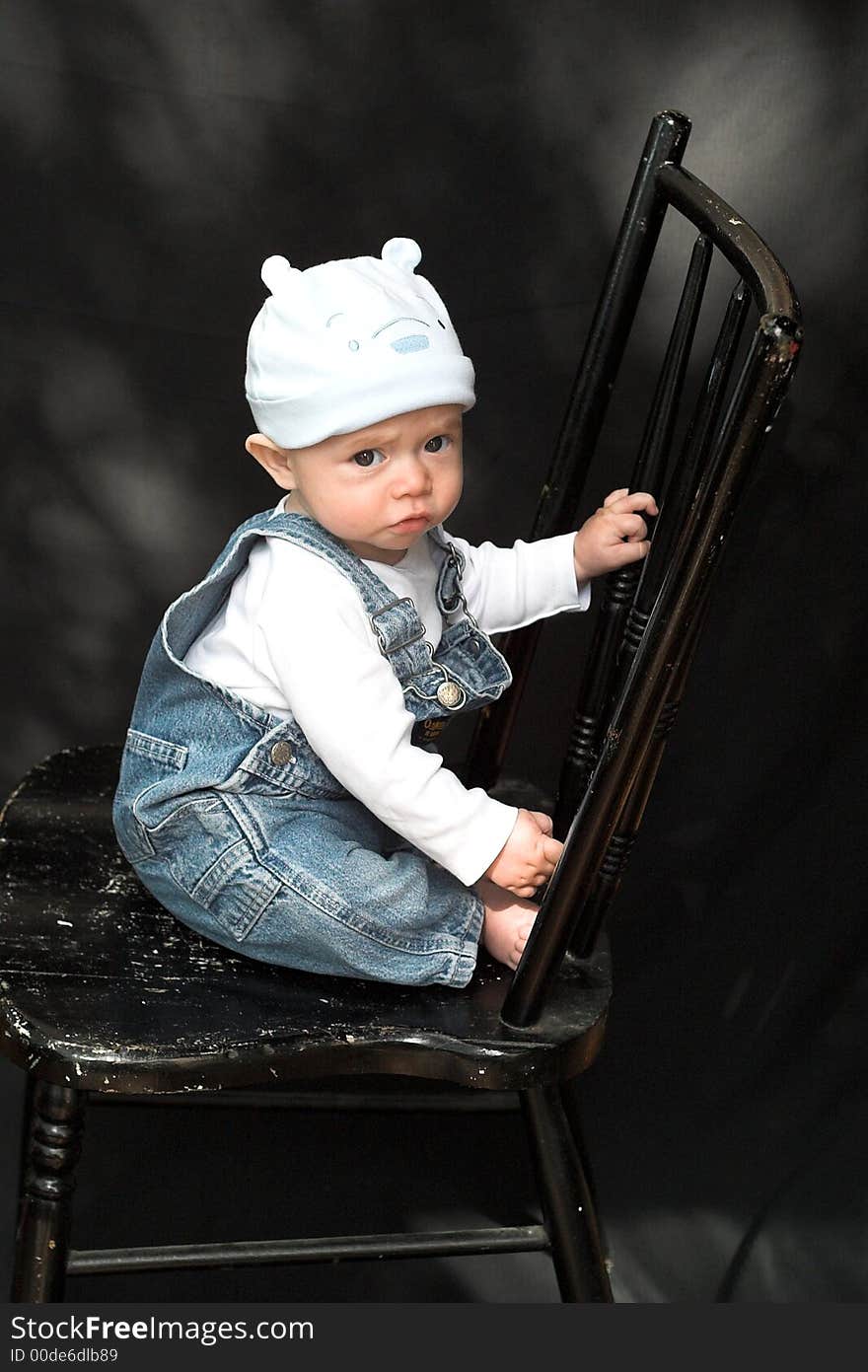 Image of adorable baby wearing denim overalls sitting on a black chair. Image of adorable baby wearing denim overalls sitting on a black chair