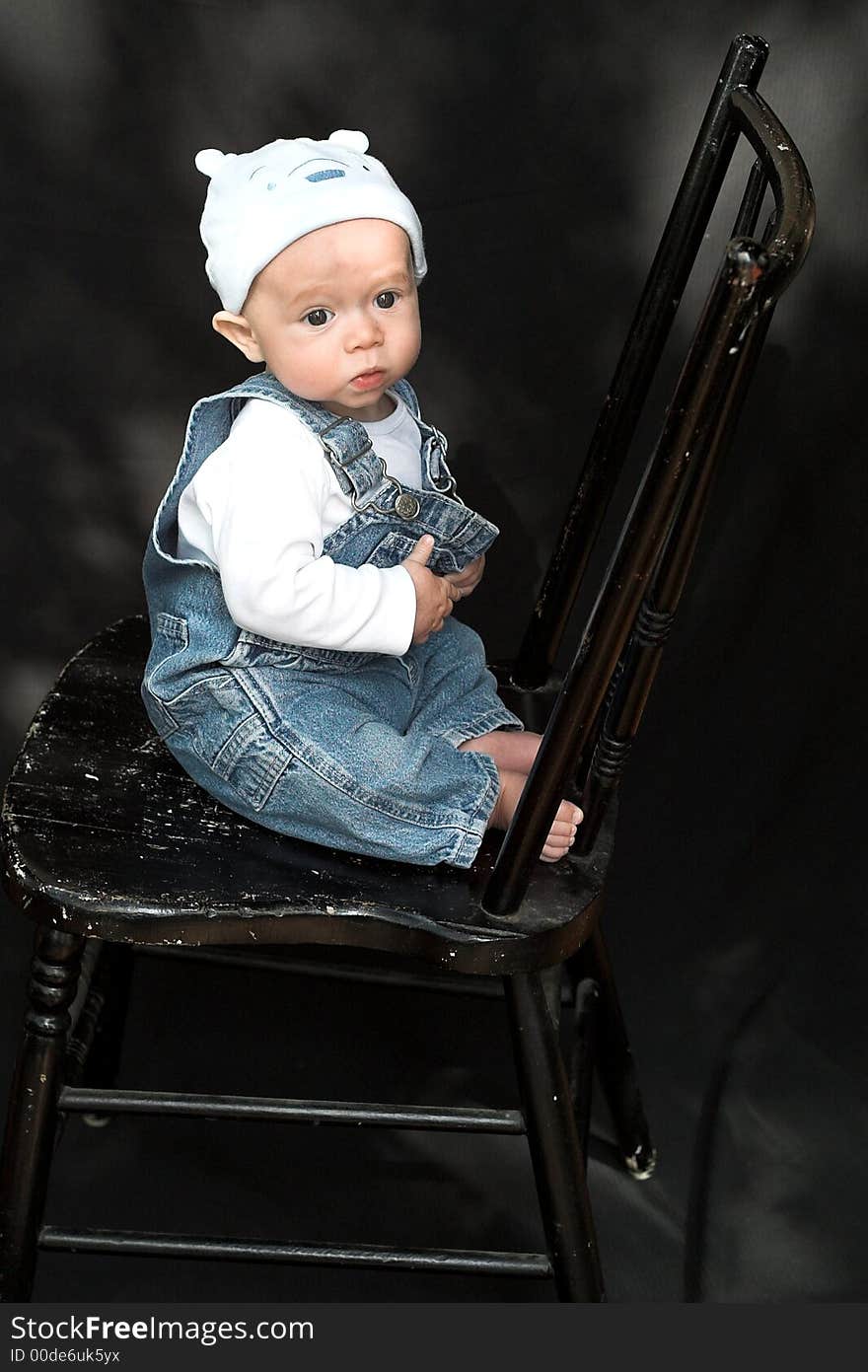 Image of adorable baby wearing denim overalls and a blue cap sitting on a black chair. Image of adorable baby wearing denim overalls and a blue cap sitting on a black chair