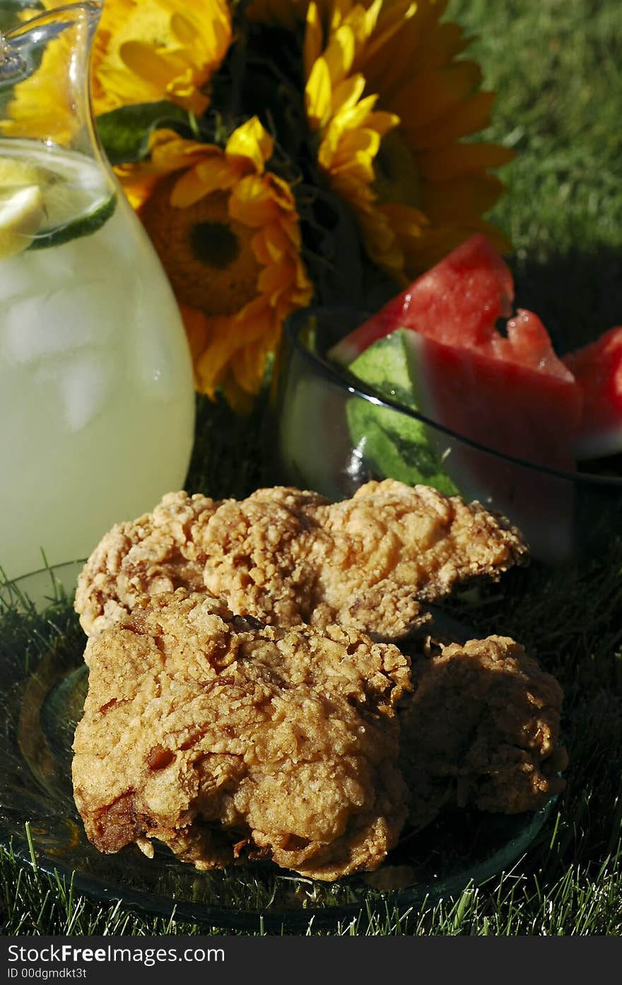 Pitcher of lemonade, plate of chicken, bowl of watermelon, vase of sunflowers on grass in summer afternoon sunlight. Pitcher of lemonade, plate of chicken, bowl of watermelon, vase of sunflowers on grass in summer afternoon sunlight