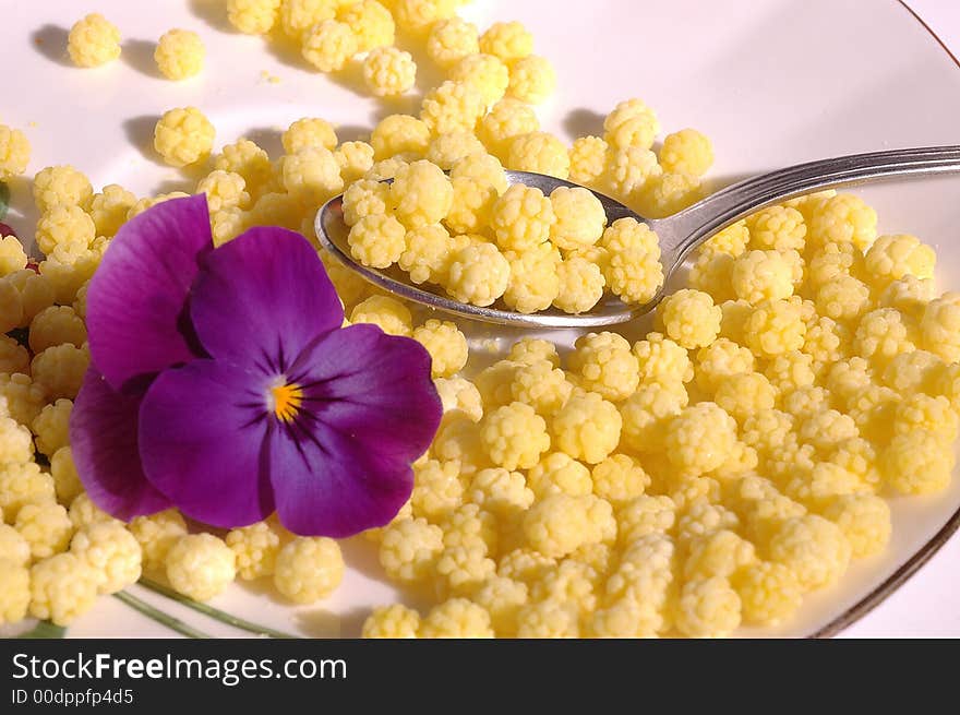 Small yellow candies like pollen with purple pansy an teaspoon on a saucer