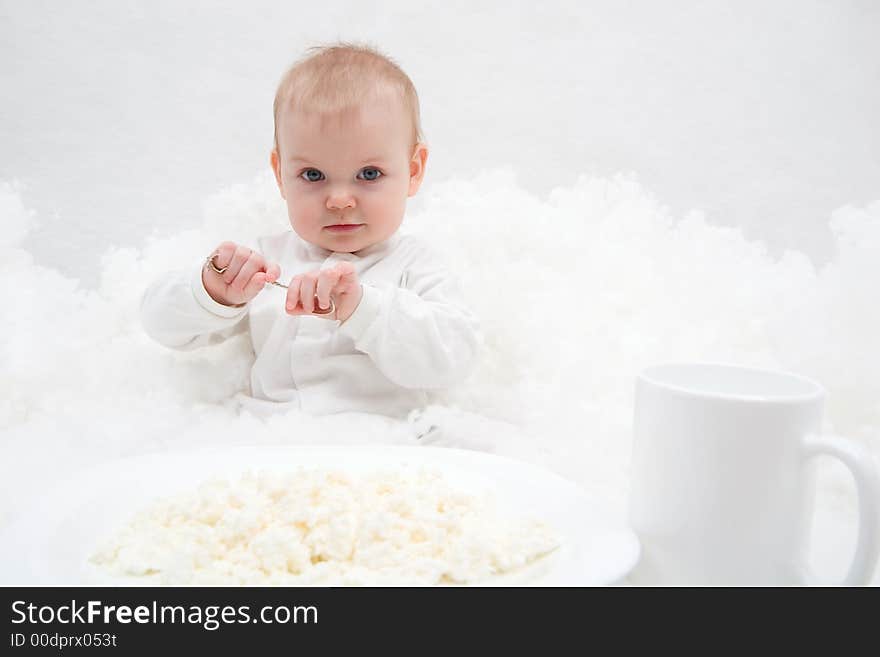 Cute little girl sitting on white blanket with spoon in her hands. There are white plate with curd and white cup in front of her. Soft-focused, focal point is on the girl. Cute little girl sitting on white blanket with spoon in her hands. There are white plate with curd and white cup in front of her. Soft-focused, focal point is on the girl