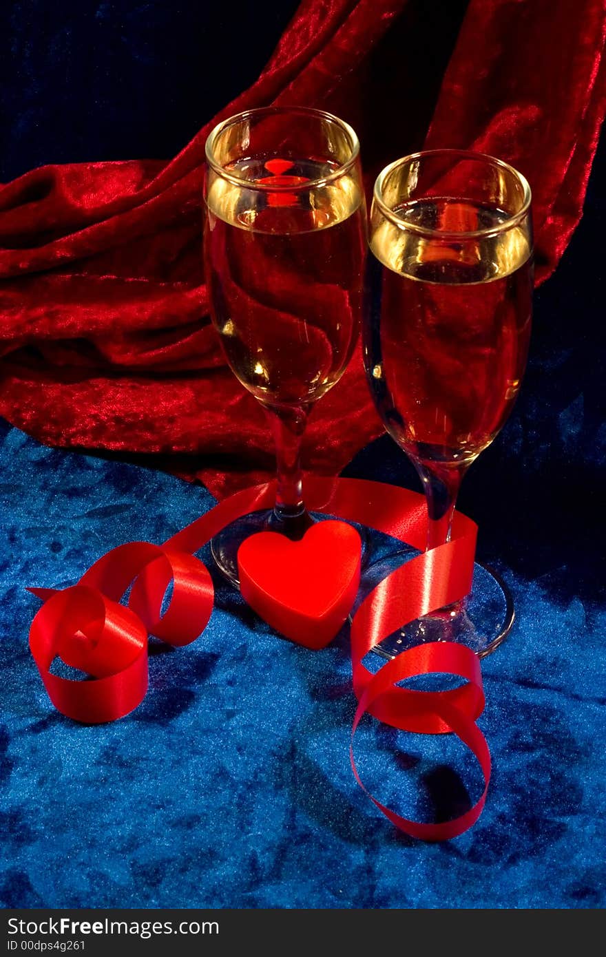 Two glasses of champagne, red twisted ribbon, and plastic heart on the blue velvet