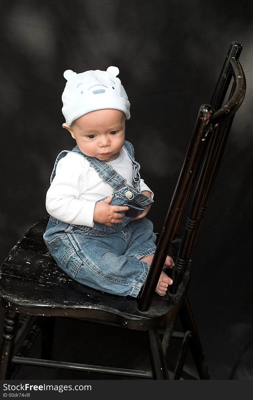 Image of adorable baby wearing denim overalls sitting on a black chair. Image of adorable baby wearing denim overalls sitting on a black chair