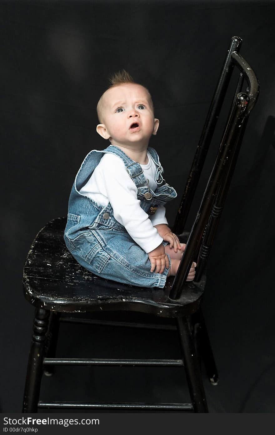 Image of adorable baby wearing denim overalls sitting on a black chair. Image of adorable baby wearing denim overalls sitting on a black chair