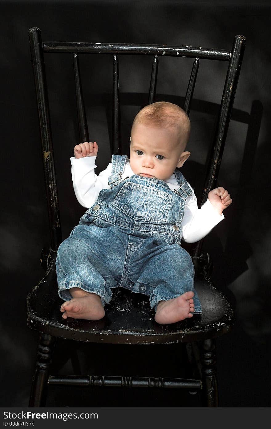 Image of adorable baby wearing denim overalls sitting on a black chair. Image of adorable baby wearing denim overalls sitting on a black chair