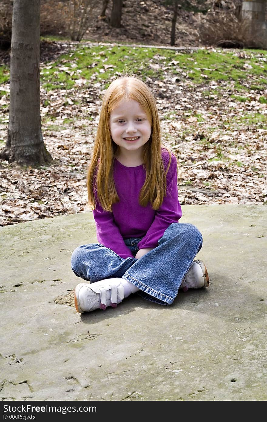 Small redhead girl sitting on a large flat stone in a park. Small redhead girl sitting on a large flat stone in a park.