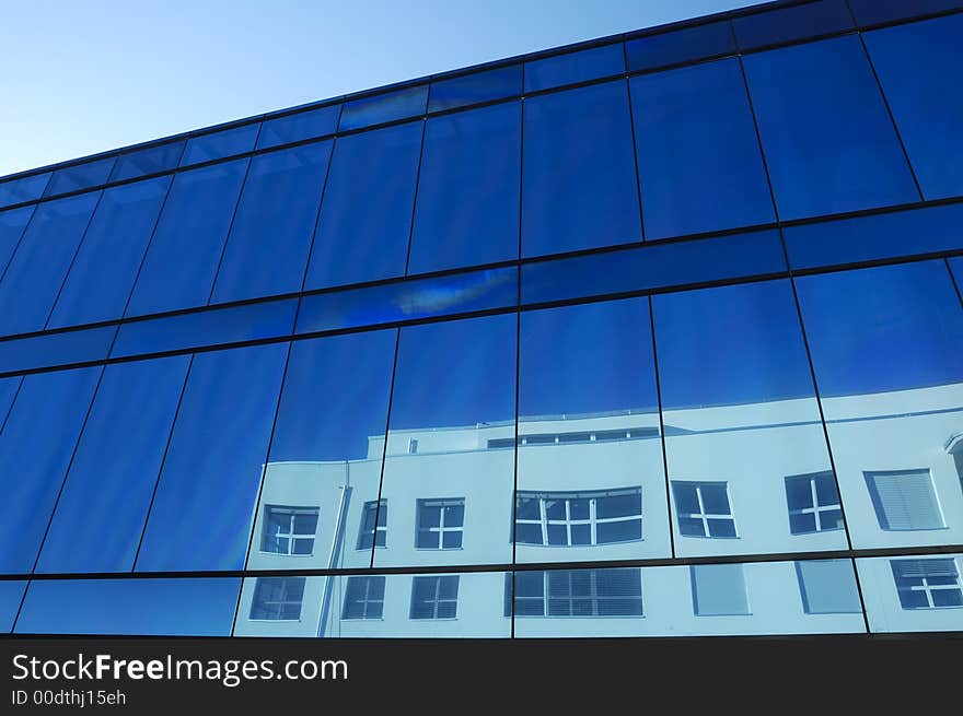 The blue glass facade of an office block with the distorted reflection of a neighbouring building showing in the windows. The blue glass facade of an office block with the distorted reflection of a neighbouring building showing in the windows.