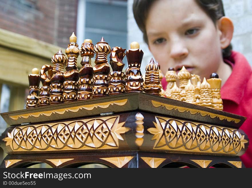 A boy peers over an ornate wooden chess set assembled for the begginning of a match. A boy peers over an ornate wooden chess set assembled for the begginning of a match.