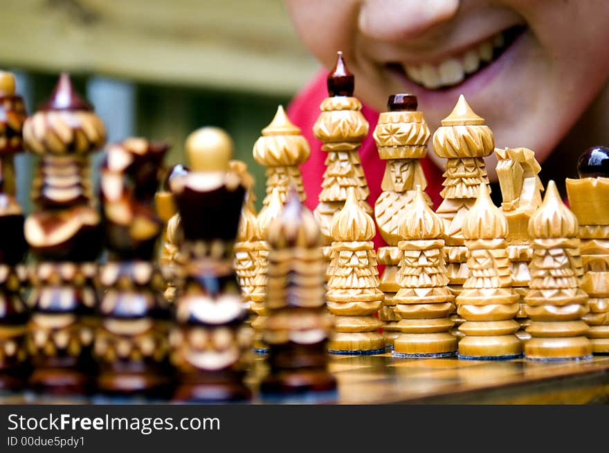 A boy's smiling mouth is visible behind the white pieces as he leans over an ornate wooden chess set assembled for the beginning of a match. A boy's smiling mouth is visible behind the white pieces as he leans over an ornate wooden chess set assembled for the beginning of a match.