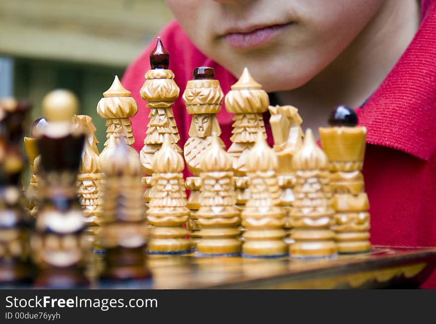 A boy's mouth with serious expression is visible behind the white pieces as he leans over an ornate wooden chess set assembled for the beginning of a match. A boy's mouth with serious expression is visible behind the white pieces as he leans over an ornate wooden chess set assembled for the beginning of a match.