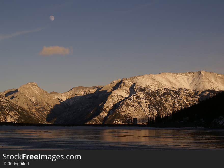 Fable & Grotto mountain at dusk in winter; Spray Lake in the foreground and moon in the background. Fable & Grotto mountain at dusk in winter; Spray Lake in the foreground and moon in the background.