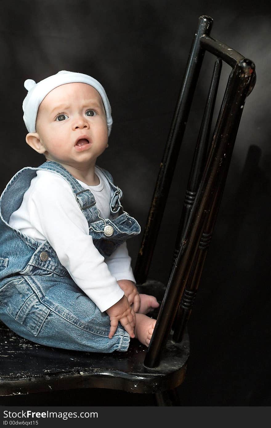 Image of adorable baby wearing denim overalls sitting on a black chair. Image of adorable baby wearing denim overalls sitting on a black chair