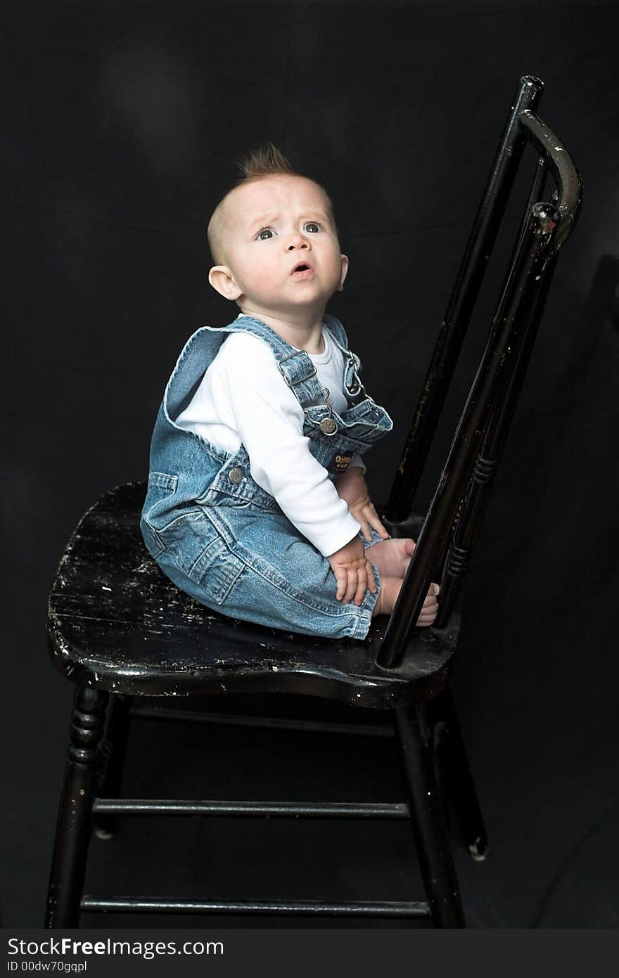 Image of adorable baby wearing denim overalls sitting on a black chair. Image of adorable baby wearing denim overalls sitting on a black chair