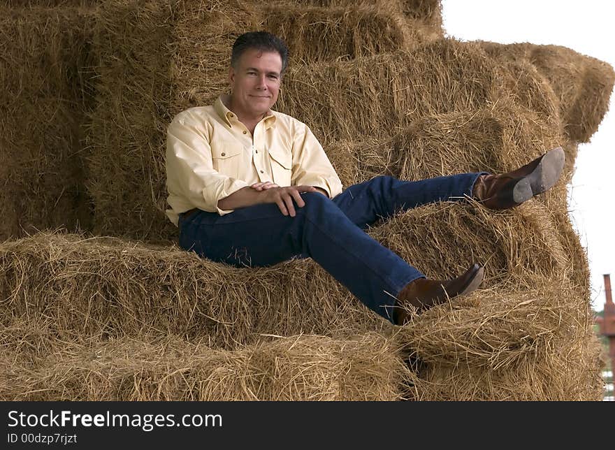 Handsome mature man sitting on a tall stack of bales of hay in the late afternoon. Handsome mature man sitting on a tall stack of bales of hay in the late afternoon.