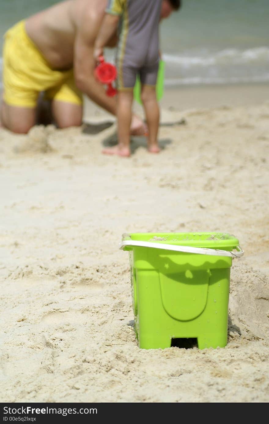 Father and son playing on beach with a green sand toy pail in the foreground. Paternal love. Father and son playing on beach with a green sand toy pail in the foreground. Paternal love.