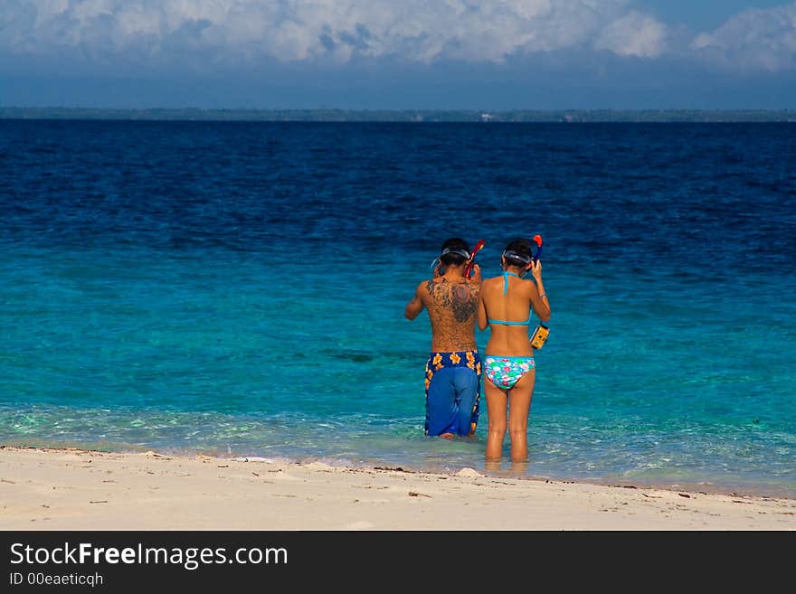 Two snorkelers prepare to swim in blue crystalline water beside a white tropical beach. Two snorkelers prepare to swim in blue crystalline water beside a white tropical beach