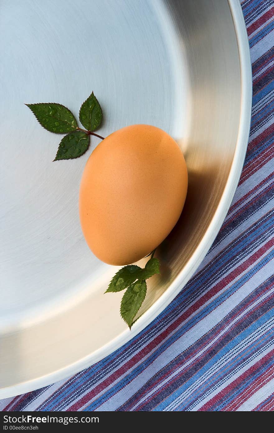 Chicken's egg in the shell in a silver frying pan. The image is shot from above. The pan is on a striped red, blue and white tablecloth. There are miniature rose leaves placed next to the egg. Chicken's egg in the shell in a silver frying pan. The image is shot from above. The pan is on a striped red, blue and white tablecloth. There are miniature rose leaves placed next to the egg