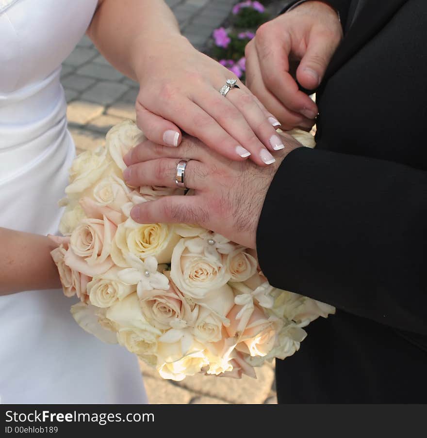 Detail of bride and groom's hands laid over bouquet of roses displaying wedding ring set. Detail of bride and groom's hands laid over bouquet of roses displaying wedding ring set