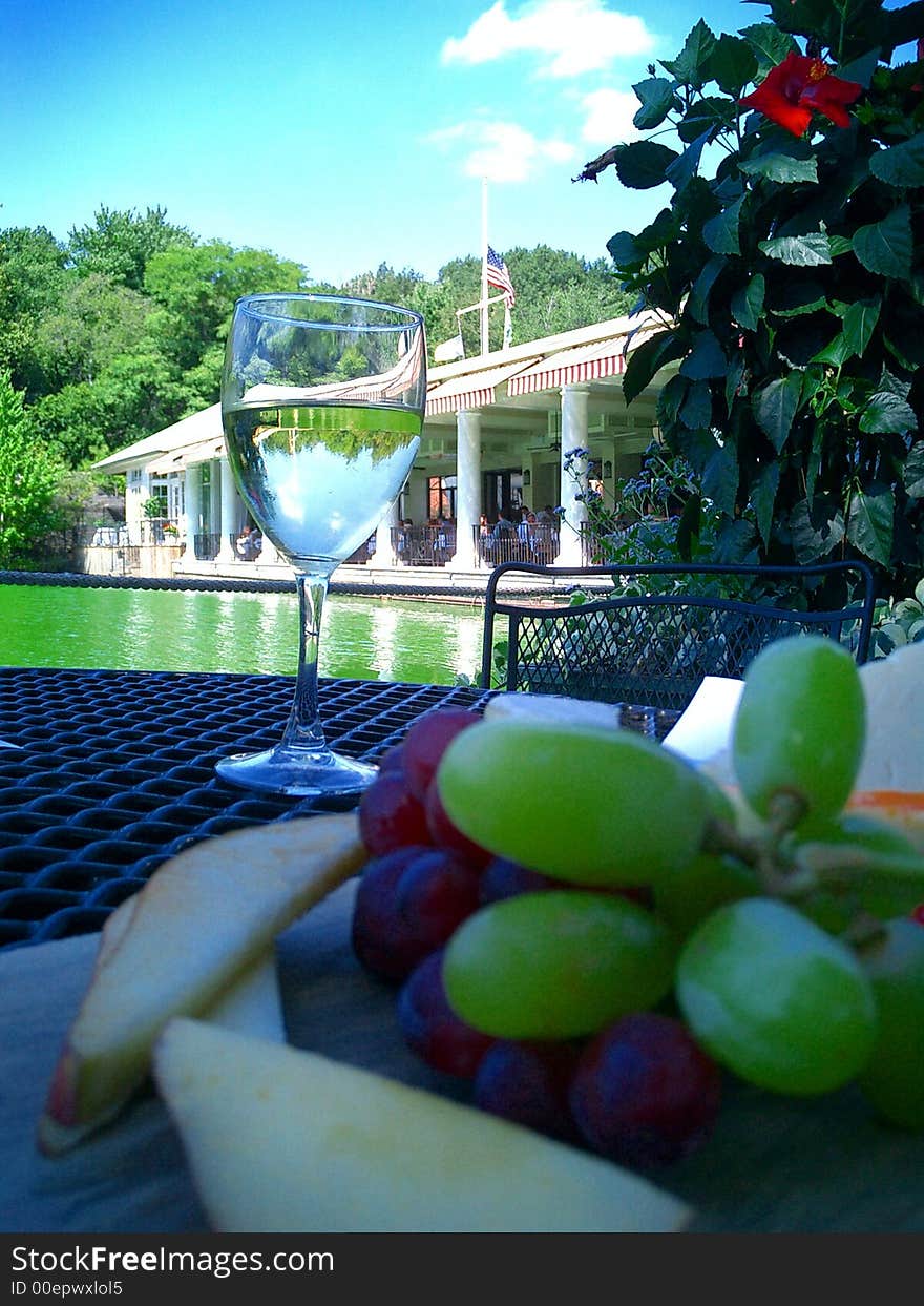 Fruit and cheese platter and glass of white wine arranged on table of restaurant terrace by a green lake with restaurant building in the background on a beautiful summer’s day. Fruit and cheese platter and glass of white wine arranged on table of restaurant terrace by a green lake with restaurant building in the background on a beautiful summer’s day