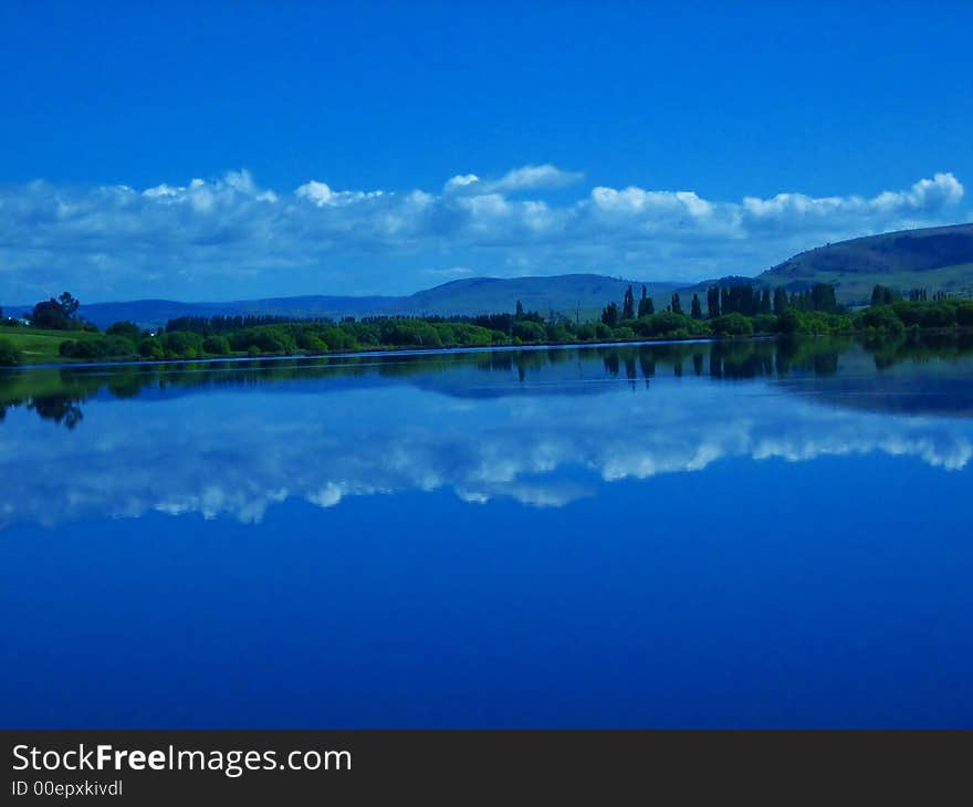 Beautiful blue sky with a few clouds, trees und hill mirrored in calm lake surface. Beautiful blue sky with a few clouds, trees und hill mirrored in calm lake surface