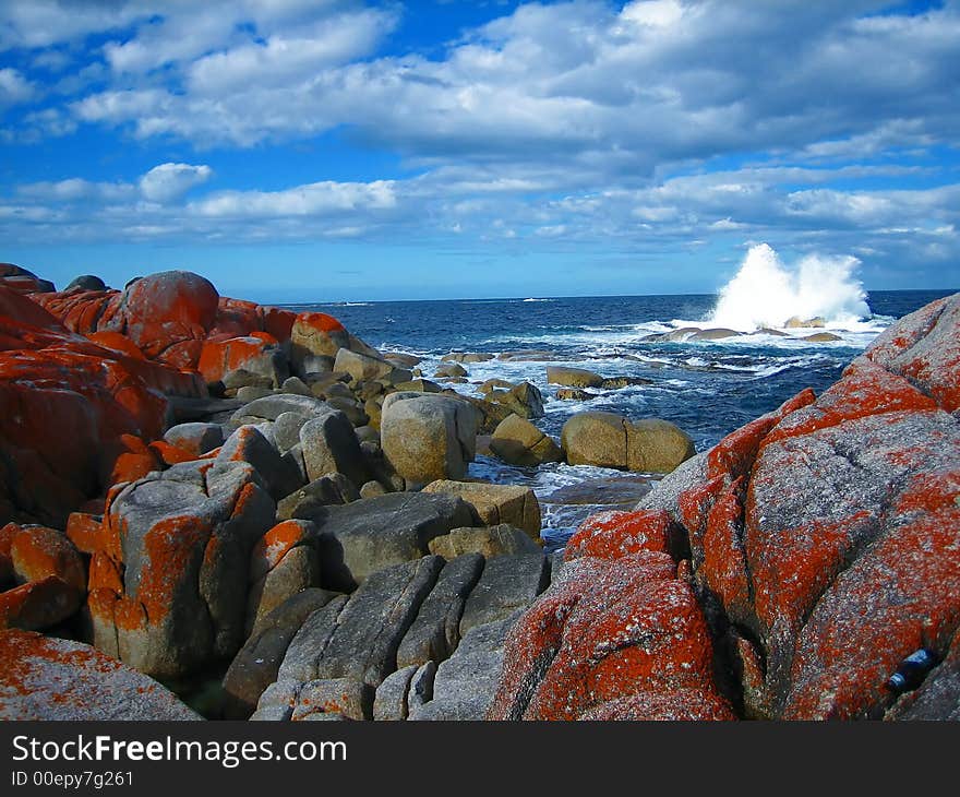 Wave splashing against rocks at coast, with boulders overgrown with orange lichen in foreground and blue sky above