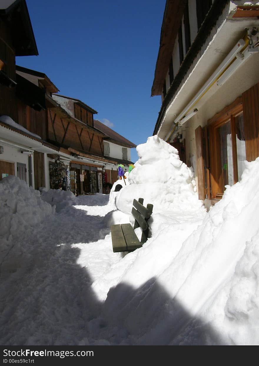 Traditional alpine village street in the snow; sunny side of the street with a wooden bench and a snow sculpture of a lion; Location: Monte Lussari in Tarvisiano; Friuli-Venezia-Giulia (FVG) region, Italy, Europe