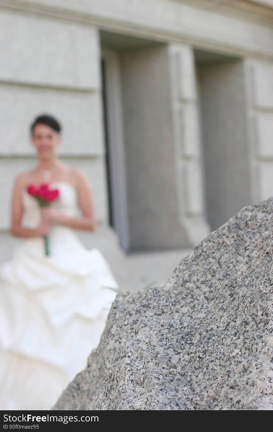 Bride Holding her bouquet standing in front of a stone building with the focus on the foreground of a large stone. Bride Holding her bouquet standing in front of a stone building with the focus on the foreground of a large stone