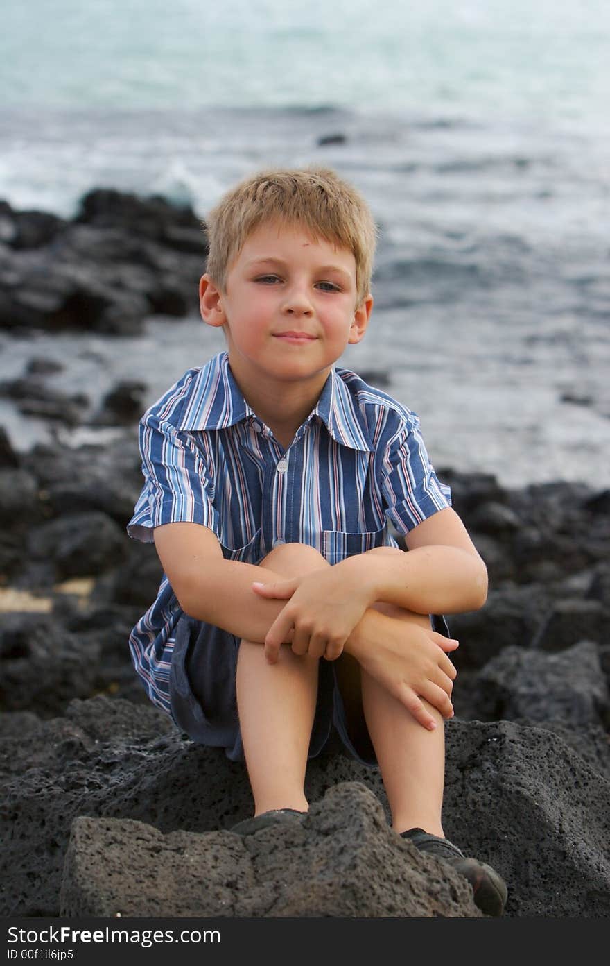 Little boy dreaming near the ocean, sitting on lava stones