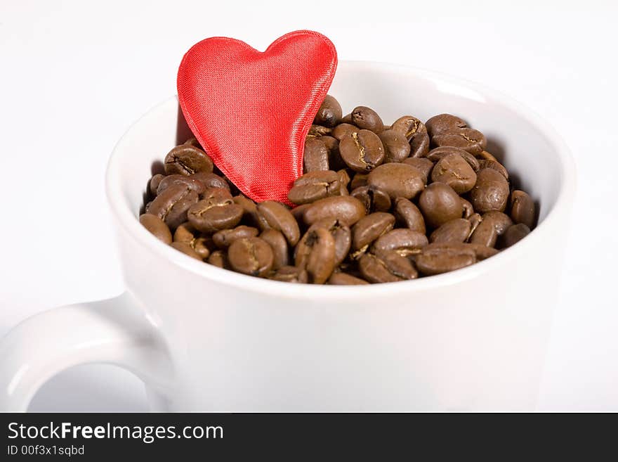 White Cup filled with coffeebeans on white background. Red heart on the beans. White Cup filled with coffeebeans on white background. Red heart on the beans.