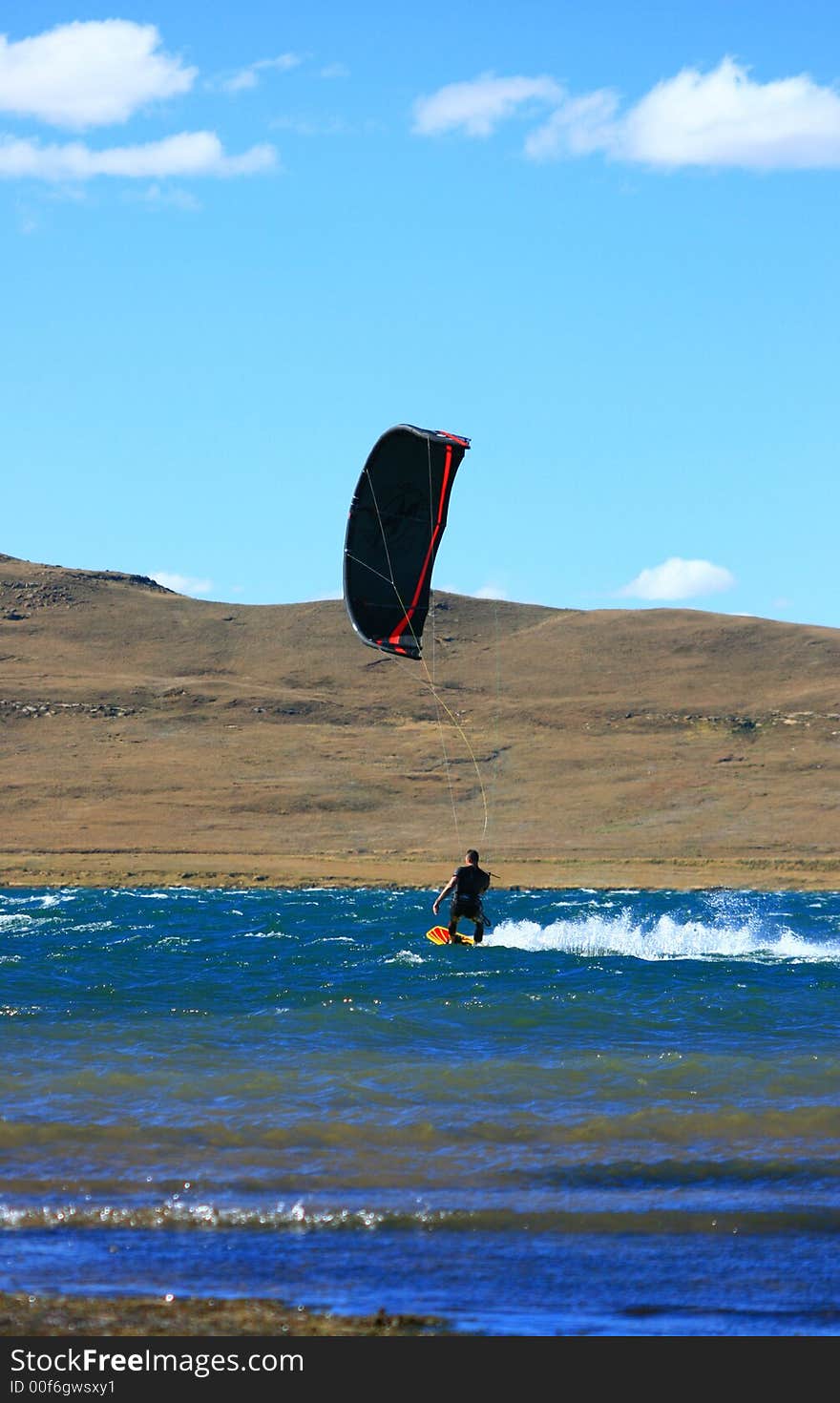 Black and red kitesurfer cruising on water at scenic dam with mountain behind, blue sky and fress water