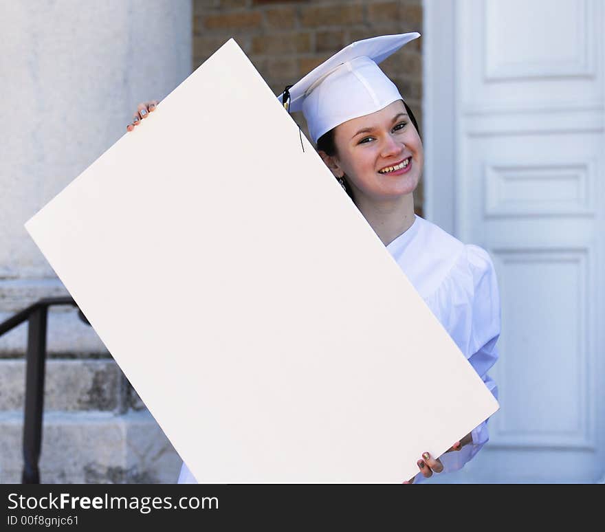 Beautiful graduate in white cap and gown holding a blank sign in front of her school. Beautiful graduate in white cap and gown holding a blank sign in front of her school.