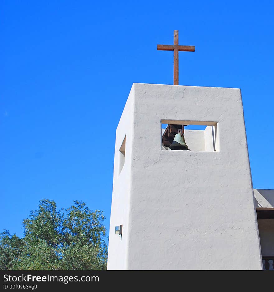 White stucco church bell tower with wooden cross atop against a blue sky