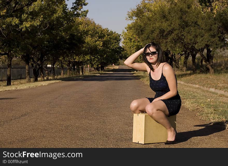 Brunette waiting for a lift sitting on suitcase roadside with hand in hair. Brunette waiting for a lift sitting on suitcase roadside with hand in hair