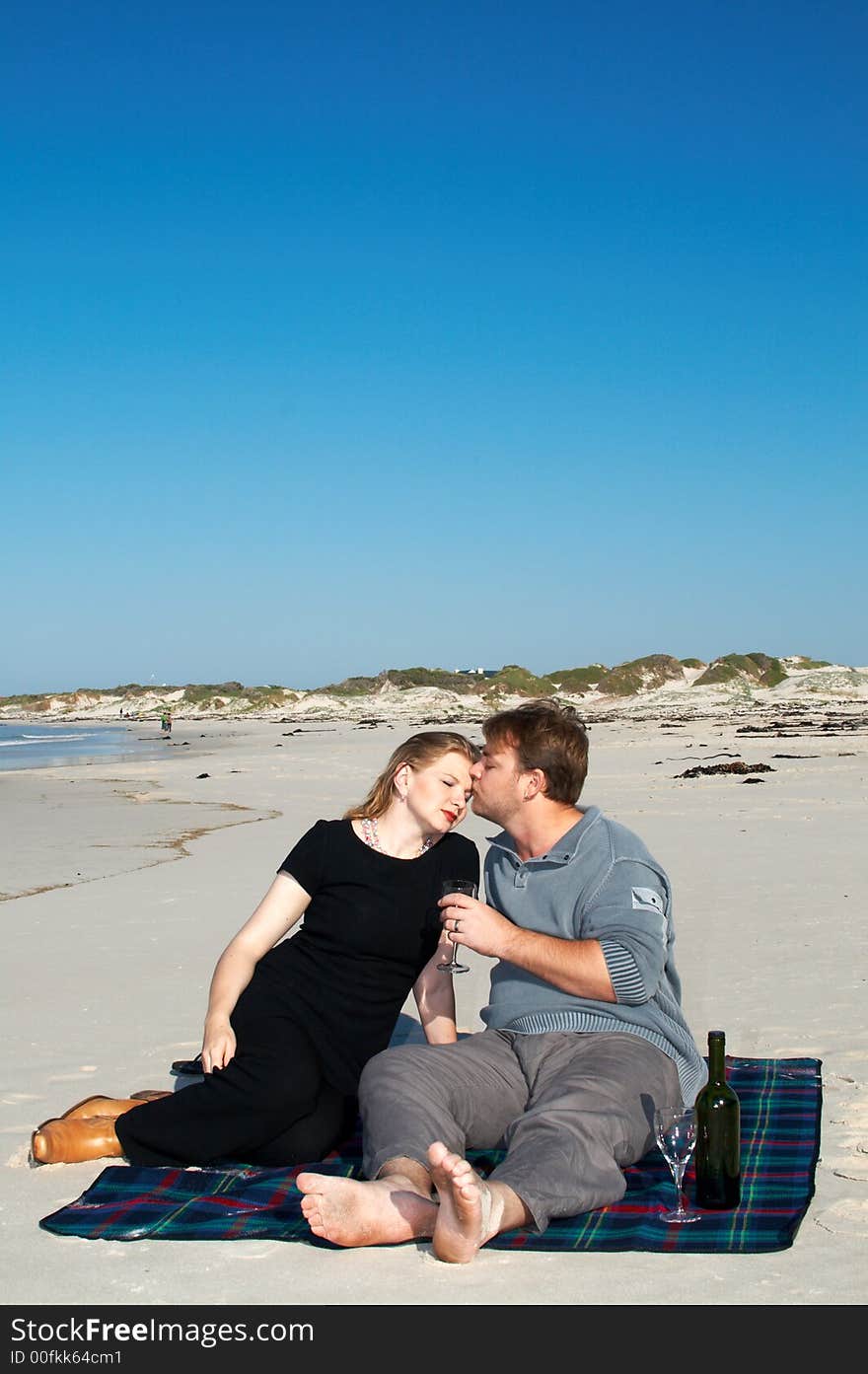 Young married couple sitting on the white sandy beach on their winter vacation in South Africa. They are enjoying the wine under the blue sky.